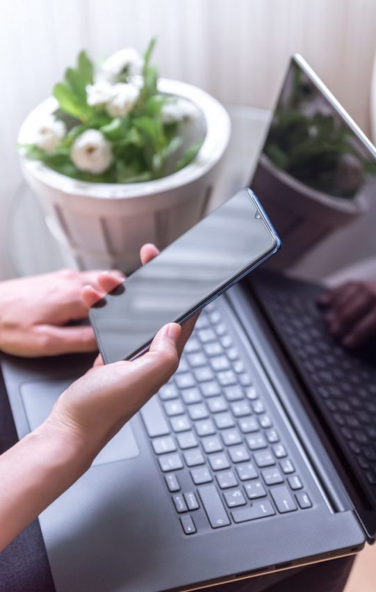 Vertical shot of a female hand holding a smartphone and laptop